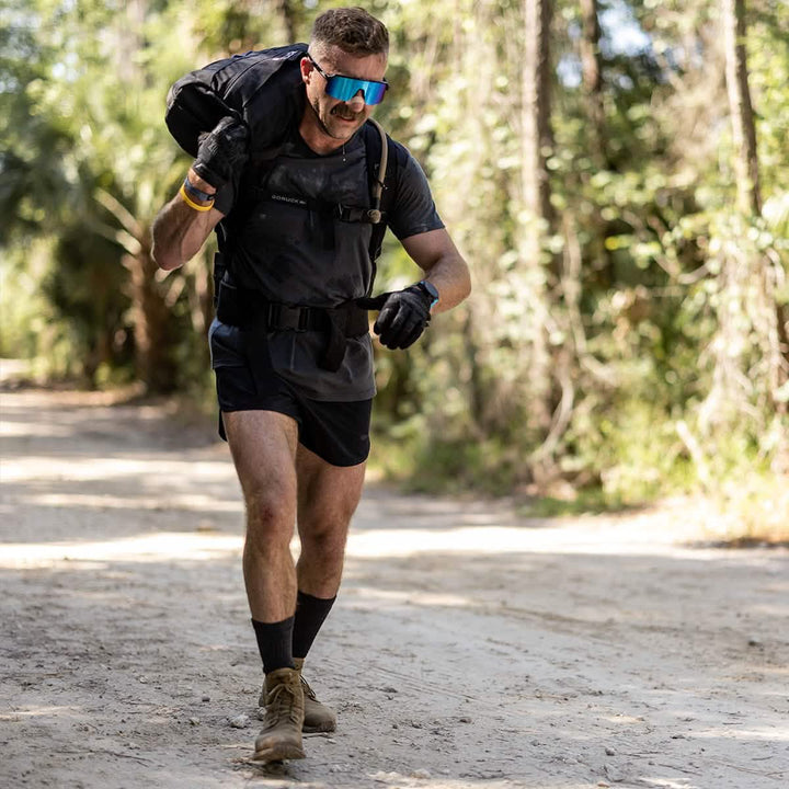 A man wearing sunglasses and GORUCK Men’s Ranger Panties - ToughStretch carries a heavy bag on his shoulder along a dirt path in a forested area. He appears focused, dressed in athletic gear made of ToughStretch fabric, including gloves and hiking boots, with tall trees providing shade in the background.