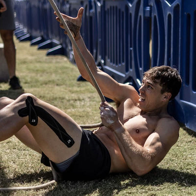 A man is shirtless, wearing black Men’s Ranger Panties - ToughStretch by GORUCK, and lying on the grass. He grips a rope with both hands, looking focused and exerting effort. The durable ToughStretch fabric withstands the challenge. A blue fence provides the backdrop to his intense workout session.