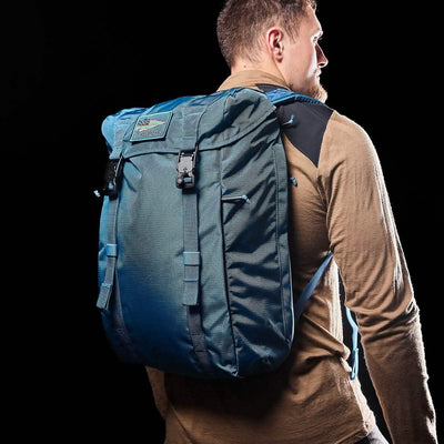 A man wearing a long-sleeve shirt in a warm, earthy tone carries the GORUCK M23 - Ripstop ROBIC® backpack in vibrant Seaspray on his back. The solid black background highlights the backpack's robust design and organizational features, making both the man and his stylish gear stand out.