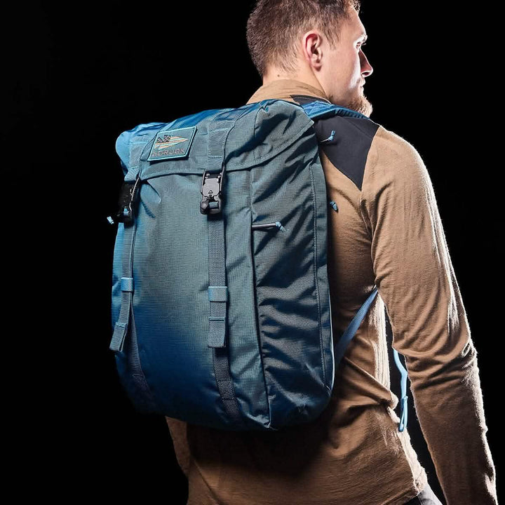 A man wearing a long-sleeve shirt in a warm, earthy tone carries the GORUCK M23 - Ripstop ROBIC® backpack in vibrant Seaspray on his back. The solid black background highlights the backpack's robust design and organizational features, making both the man and his stylish gear stand out.