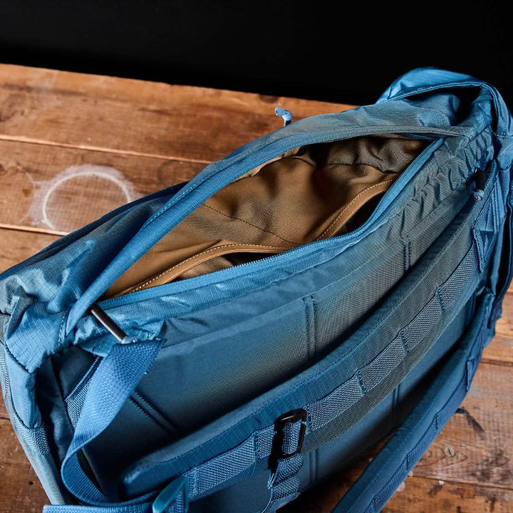 The M23 Ripstop ROBIC® by GORUCK in Seaspray + Speed Tan features a modern design with a partially open top zipper, revealing an interior of brown lining crafted from durable materials. Multiple straps and buckles indicate its functionality for travel or outdoor activities, all while it rests on a rustic wooden surface, making it perfect for meticulous organization.