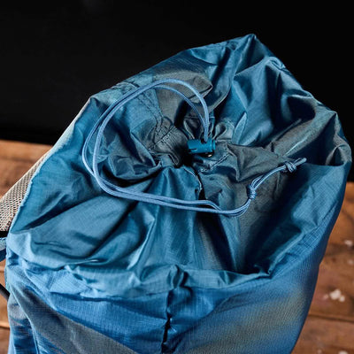 The M23 - Ripstop ROBIC® rucksack from GORUCK, in Seaspray and Speed Tan, is open, showcasing its organized interior. Made with durable materials, the drawstring closure is looped through a toggle for secure fastening. The backpack sits on a wooden surface against a black background.