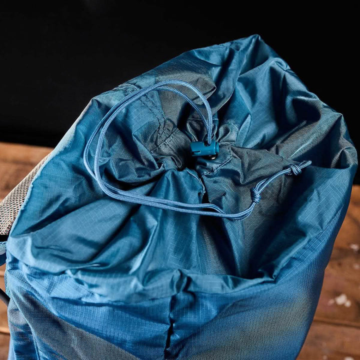 The M23 - Ripstop ROBIC® rucksack from GORUCK, in Seaspray and Speed Tan, is open, showcasing its organized interior. Made with durable materials, the drawstring closure is looped through a toggle for secure fastening. The backpack sits on a wooden surface against a black background.