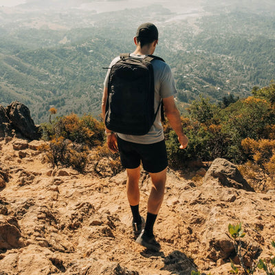 A person wearing a cap, T-shirt, shorts, and a pair of GORUCK's Mackall - Black + Forged Iron shoes is hiking down a rocky trail. They overlook a scenic view of a town surrounded by trees and hills under a clear sky. The trek feels like Special Forces training, pushing them beyond the ordinary with each step.