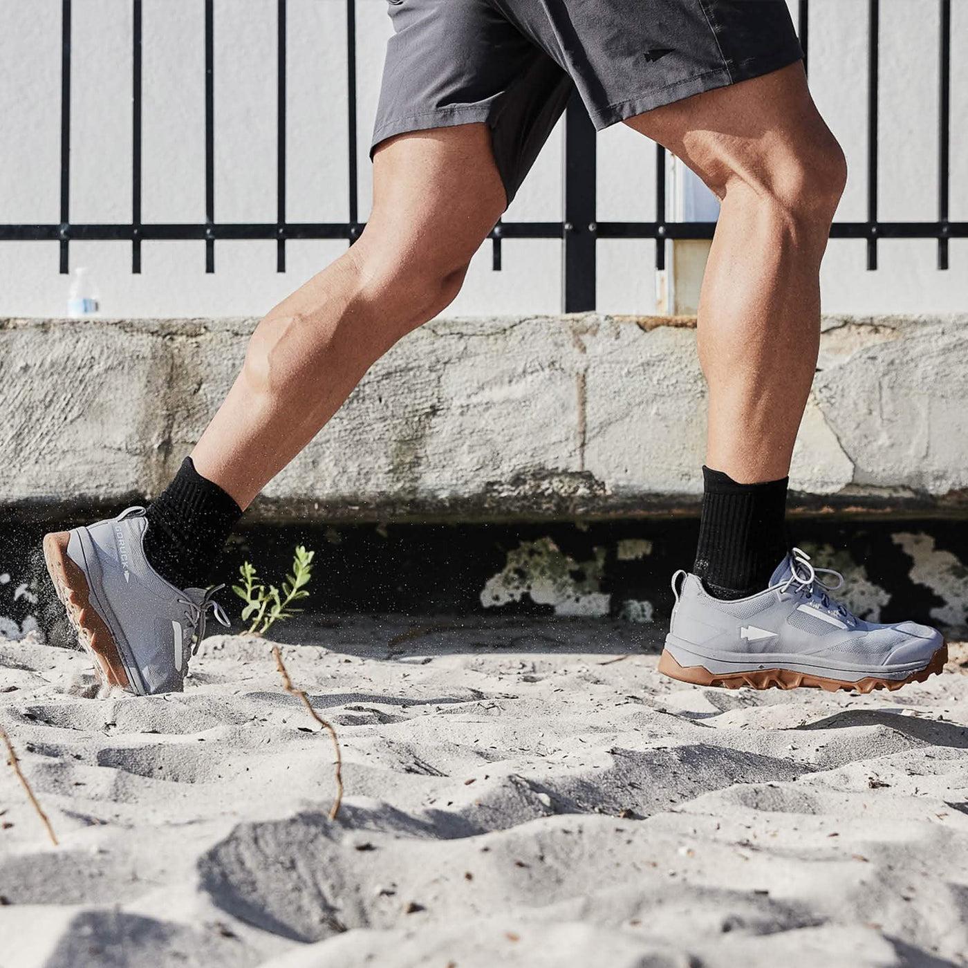 While jogging on a sandy surface, the person is wearing gray GORUCK trail shoes named Mackall - Chiseled Stone + Gum, known for their triple compound outsole. They are paired with black socks and gray shorts. The background includes a stone wall and a black metal fence.