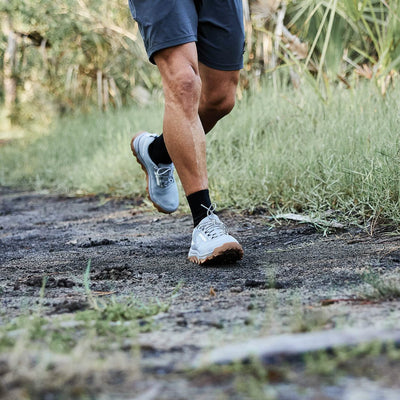 A person wearing gray Mackall shorts and GORUCK trail shoes with a triple compound outsole jogs on a dirt trail surrounded by grass and greenery. The focus is on their legs and feet, suggesting outdoor exercise.