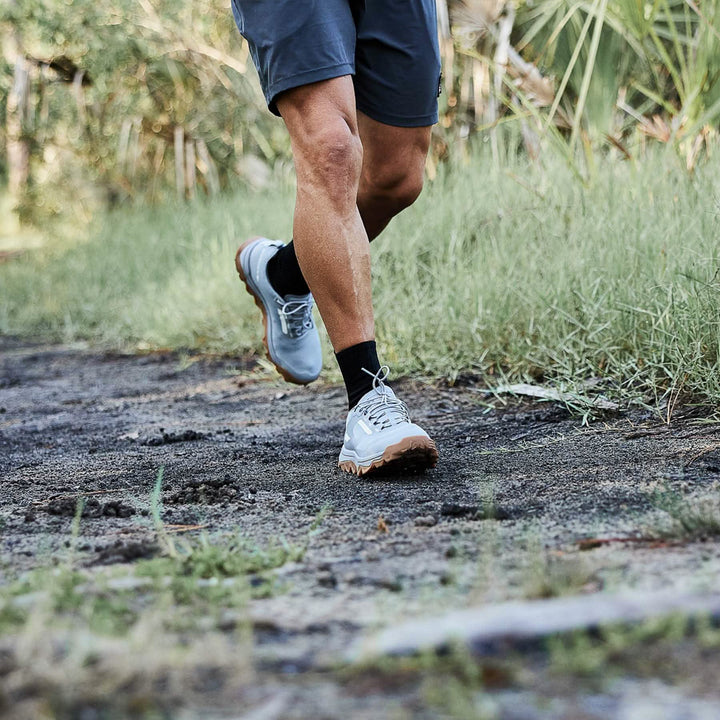 A person wearing gray Mackall shorts and GORUCK trail shoes with a triple compound outsole jogs on a dirt trail surrounded by grass and greenery. The focus is on their legs and feet, suggesting outdoor exercise.