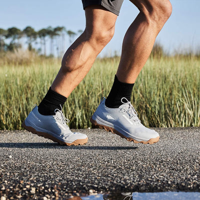 A person running on a pathway near a grassy field is sporting Mackall - Chiseled Stone + Gum trail running shoes by GORUCK, featuring a triple compound outsole. They are complemented by black socks and gray shorts, against the serene backdrop of tall grass and blurred trees under the clear sky.
