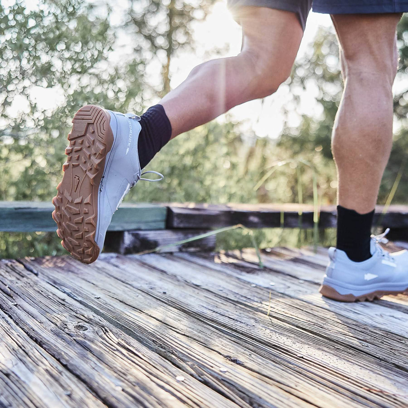 A jogger wearing the Mackall - Chiseled Stone + Gum trail shoes from GORUCK moves along a wooden boardwalk, with sunlight filtering through the trees. The emphasis is on their legs and footwear, indicating an outdoor run in a natural setting enhanced by the triple compound outsole for superior traction and support.