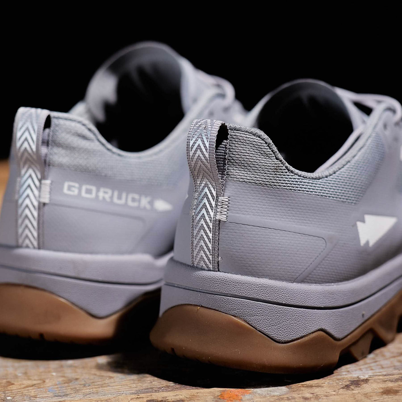 A close-up of the GORUCK Mackall trail shoes in Chiseled Stone + Gum placed on a wooden surface showcases their brown outsoles, with the brand name proudly visible on the back. The shoes feature white chevron patterns near the heel tabs, adding a touch of style to their rugged design.