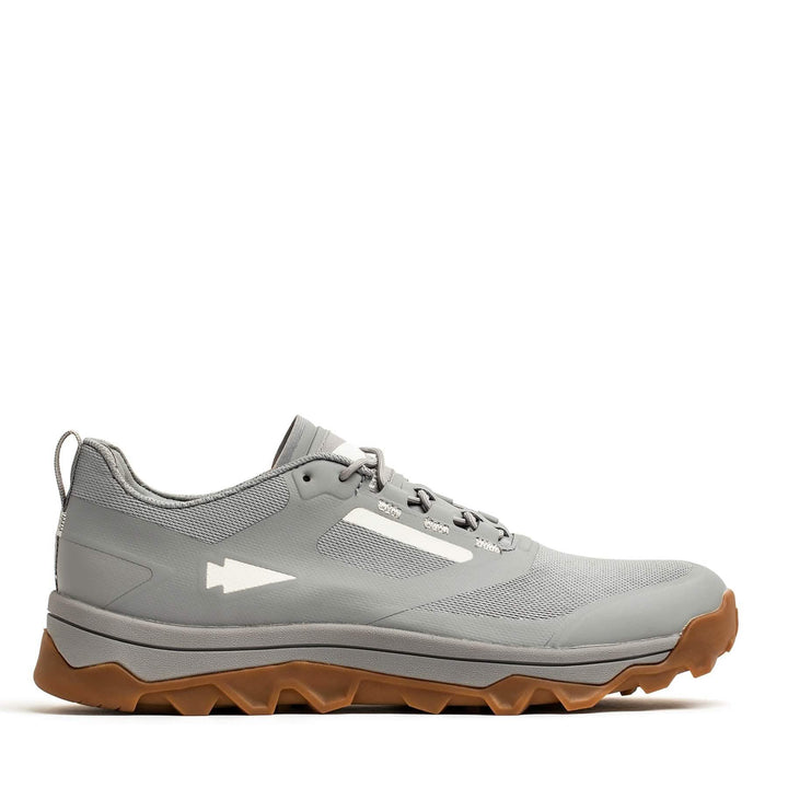 The Mackall - Chiseled Stone + Gum by GORUCK is a rucking shoe in gray with a rugged, textured sole and a white arrow logo on the side, all set against a plain white background. Its brown triple compound outsole provides outstanding contrast and grip, making it ideal for any trail adventure.