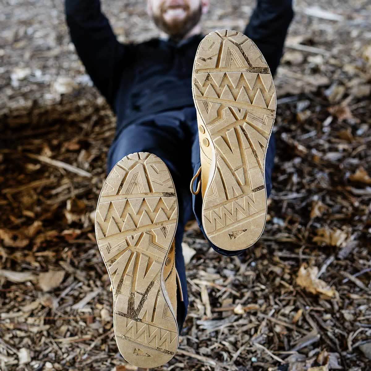 Dressed in dark clothing, an individual is lying on the ground with a smile, their feet positioned near the camera. The shoes they are wearing showcase the distinctive MACV-1 - Mid Top by GORUCK in Coyote Suede, featuring a characteristic zig-zag tread pattern and an all-terrain outsole as they rest on a bed of leaves and bark.
