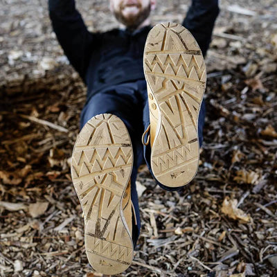 Dressed in dark clothing, an individual is lying on the ground with a smile, their feet positioned near the camera. The shoes they are wearing showcase the distinctive MACV-1 - Mid Top by GORUCK in Coyote Suede, featuring a characteristic zig-zag tread pattern and an all-terrain outsole as they rest on a bed of leaves and bark.