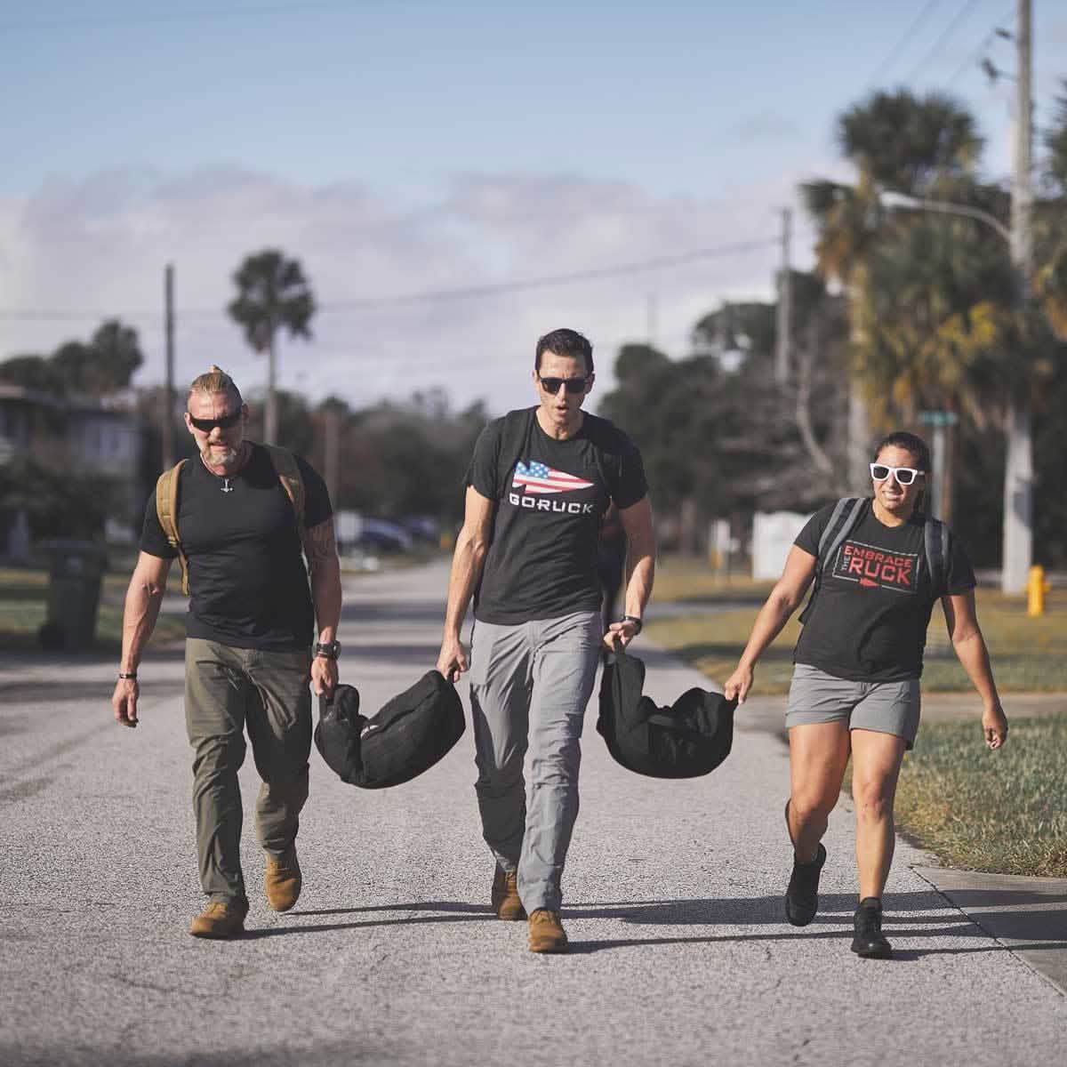 Wearing GORUCK MACV-1 - Mid Top - Coyote Suede shoes, three people walk along a suburban street with weighted bags. Dressed in casual athletic wear, sunglasses, and backpacks, they enjoy the path lined with palm trees and houses. Their all-terrain outsoles provide confident footing under the partly cloudy sky.