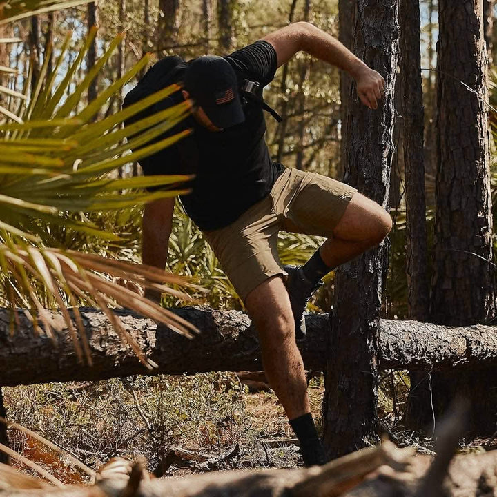 A person wearing a black shirt, khaki shorts, and a black cap is climbing over a fallen tree in a dense forest, their footing sure in GORUCK's Merino Challenge Socks - Crew. The scene is lit by sunlight filtering through the trees, casting serene patterns over this peaceful adventure.