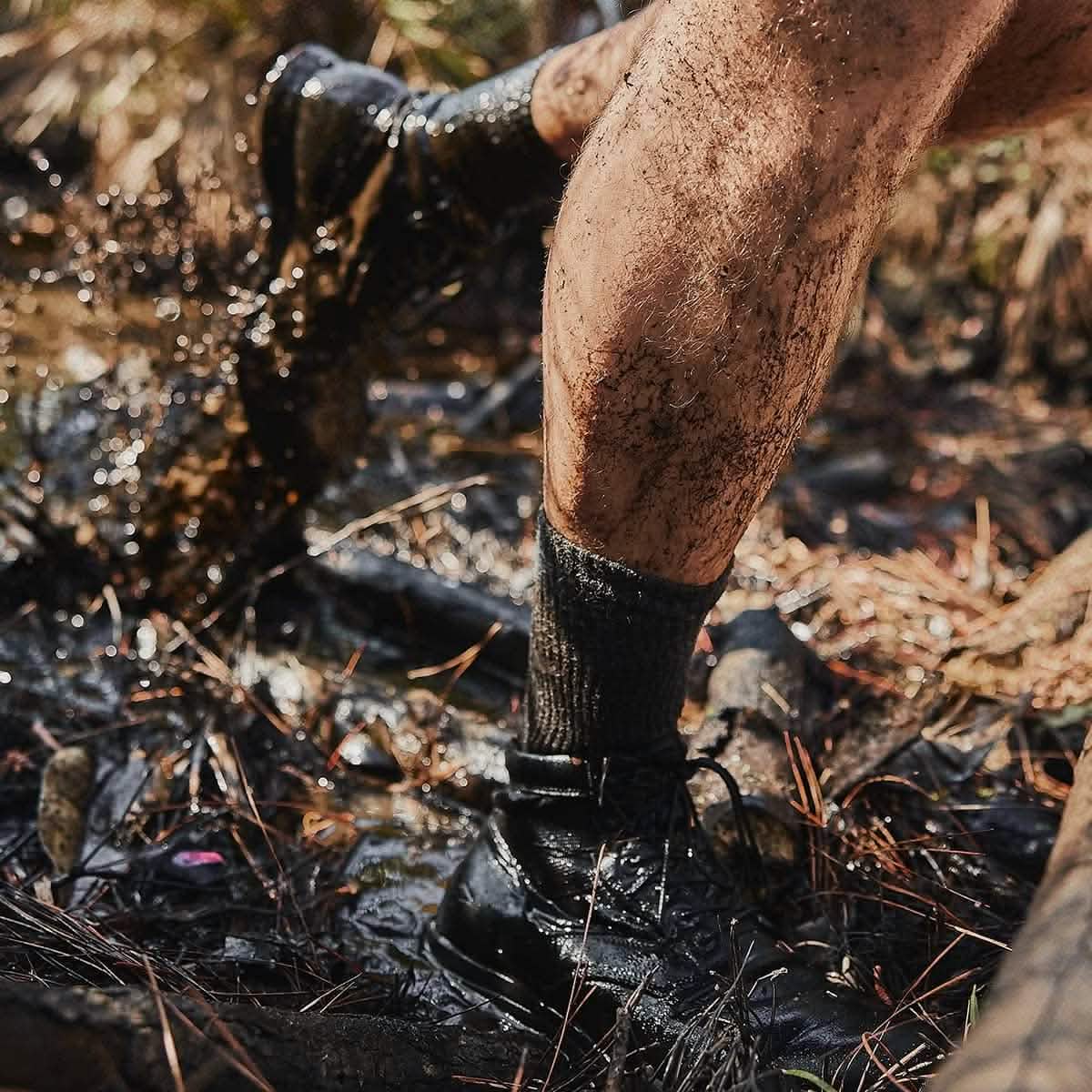Close-up of a person’s muddy legs and boots, safeguarded by GORUCK's Merino Challenge Socks - Crew, as they trek through a wet, forested terrain. The ground is blanketed with water, mud, and fallen leaves, while sunlight filters through the trees.
