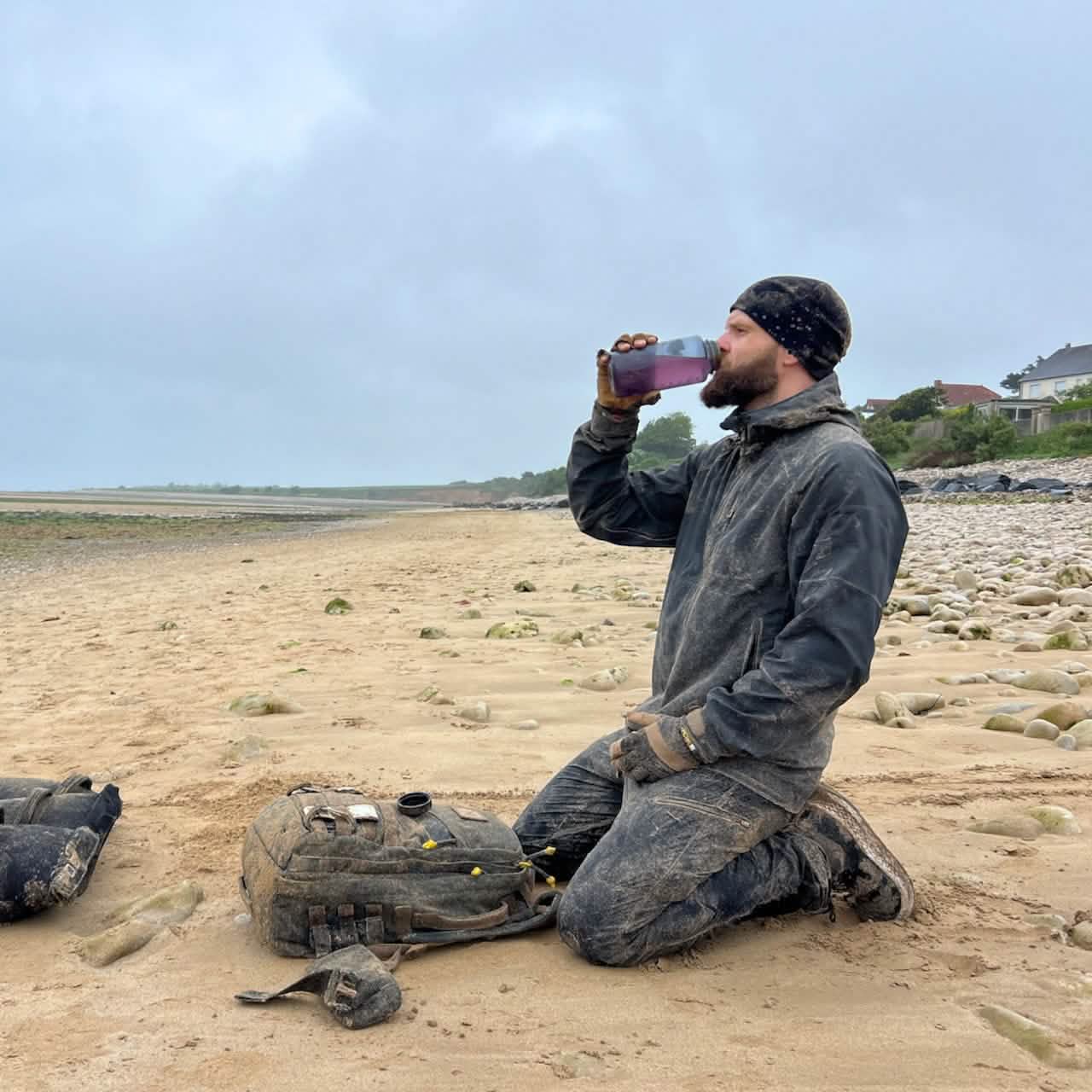 A person kneels on a beach, wearing a beanie and a muddy outfit, sipping from a 32oz Nalgene Water Bottle - GORUCK Spearhead. A backpack lies in the sand nearby, with a cloudy sky and rocky shoreline in the background.