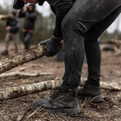 A person dressed in rugged outdoor clothing, featuring the black MACV-2 - Mid Top - Origins boots from GORUCK and gloves, is seen bending over while holding a thick branch on a forest floor. Blurred figures and trees create a wilderness setting reminiscent of Special Forces operations in the background.
