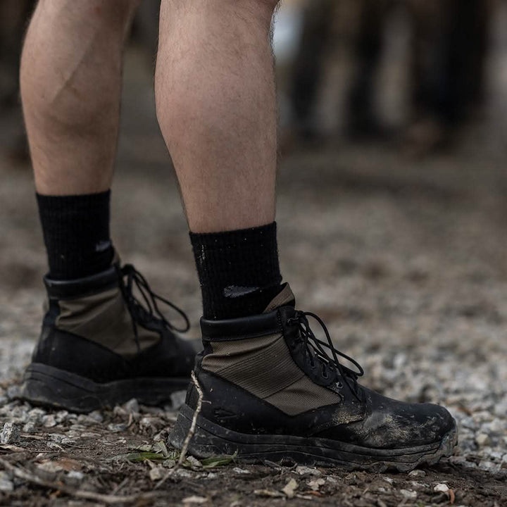 Close-up of a person's legs wearing GORUCK's MACV-2 - Mid Top - Origins on a gravel path. The background is blurred, suggesting an outdoor environment, possibly a challenging hiking trail.