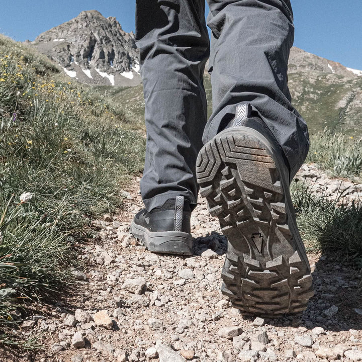 Clad in the GORUCK Mackall - Black + Forged Iron trail shoes, an individual crosses a rugged mountain path dotted with grass and wildflowers. Towering peaks rise dramatically against a clear sky, capturing the spirit of Special Forces training.