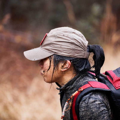 A person sporting a GORUCK Performance TAC Hat - TOUGHDRY strides through a natural setting, their long dark hair tied back and outdoor gear crafted from sweat-wicking material. The motion-blurred background emphasizes the focus on the adventurous explorer.