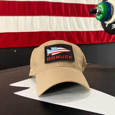 A tan Performance TAC Hat - TOUGHDRY from GORUCK, adorned with an American flag patch, rests on a black surface. Made with ToughDry fabric and featuring a VELCRO closure, it is prominently displayed against a blurred backdrop of gym equipment and a large American flag.