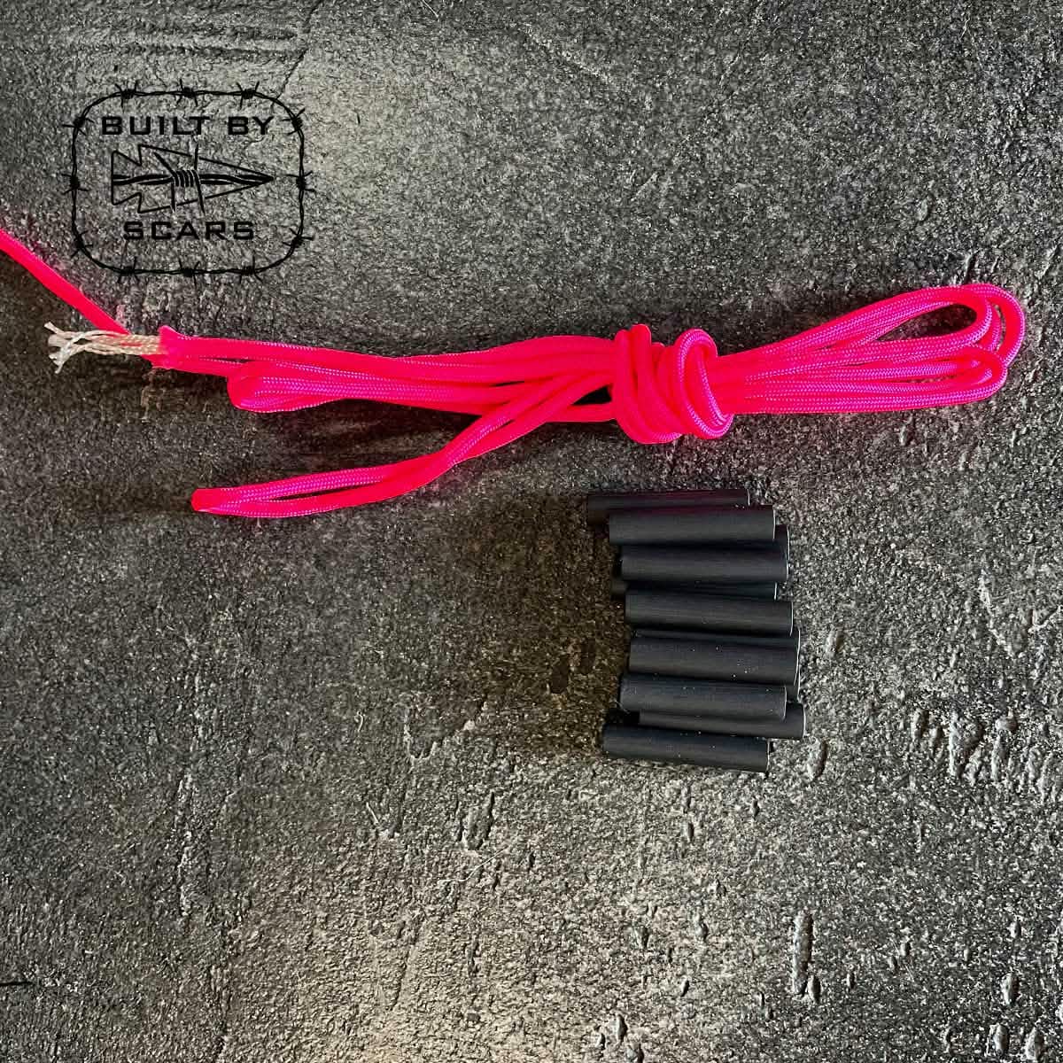 A coiled pink cord is positioned beside eight small black cylindrical pieces, resembling components of the Paracord Kit - Built By SCARS from GORUCK, arranged in a stepped pattern on a textured dark surface. The Built by SCARS logo is visible in the top left corner.