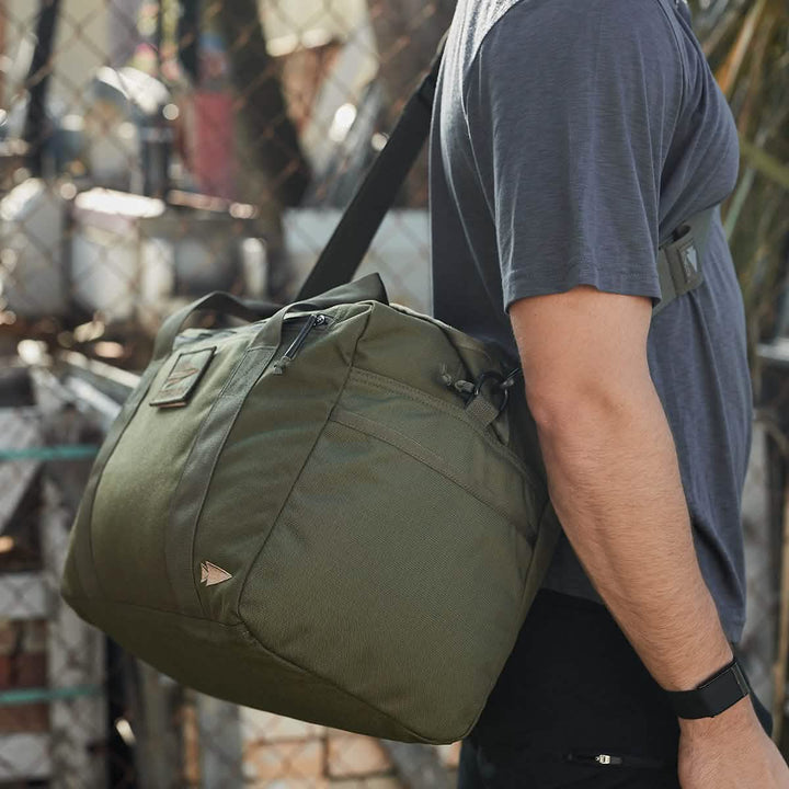 A person wearing a gray T-shirt carries a large, green Kit Bag (Includes Shoulder Strap) by GORUCK on their shoulder. The background features a metal fence and blurred outdoor elements, suggesting an urban environment.