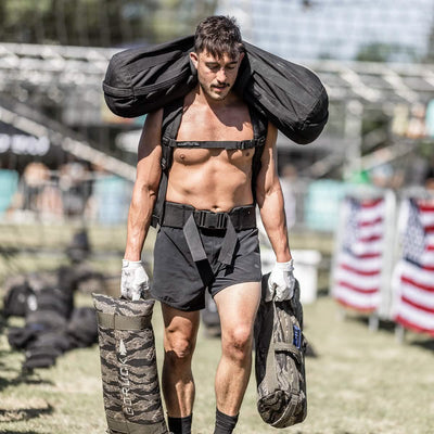A muscular man in GORUCK's Men’s Ranger Panties - ToughStretch and gloves carries heavy sandbags over his shoulders and in each hand while walking across a grassy area. American flags and obstacle course equipment are visible in the background, showcasing resilience with a Scars Lifetime Guarantee.