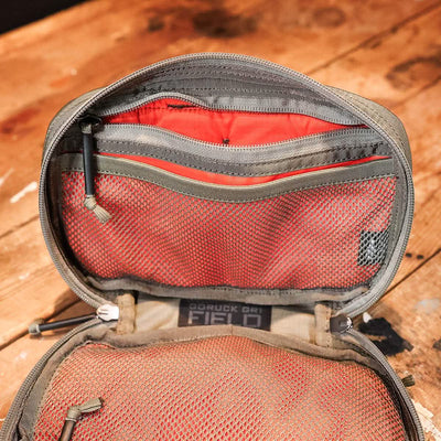 A GR1 Field Pocket - Ripstop ROBIC® by GORUCK, featuring a green and orange design crafted from Ripstop Nylon, is shown on a wooden surface. This electronics organizer includes zipper compartments and mesh pockets, showcasing its water-resistant storage capacity and detailed design.