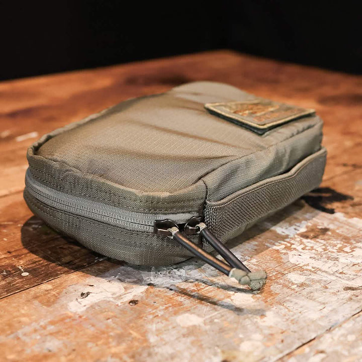 On a rustic wooden surface rests the GR1 Field Pocket, a small electronics organizer by GORUCK. Crafted from olive green Ripstop ROBIC®, this water-resistant pouch features a patch on top and two convenient zipper tabs for easy access.
