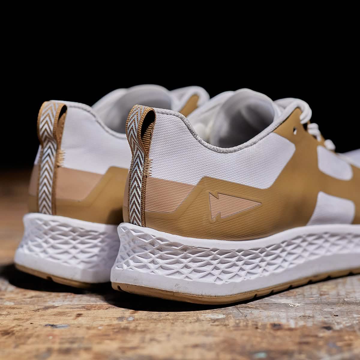 Introducing the Men's Rough Runner - Speed Tan by GORUCK: these white and tan sneakers feature textured white soles and are perfect for high mileage performance. Displayed elegantly on a wooden surface, they showcase a geometric design on the heel tab along with a minimalist arrow logo on the side, combining both style and function.