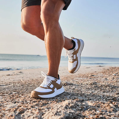 A person jogging on a sandy beach in GORUCK's Men's Rough Runner - Speed Tan sneakers embodies the essence of a Rough Runner. The ocean and clear sky frame the scene, focusing on their lower legs to highlight high mileage movement and relentless outdoor activity.