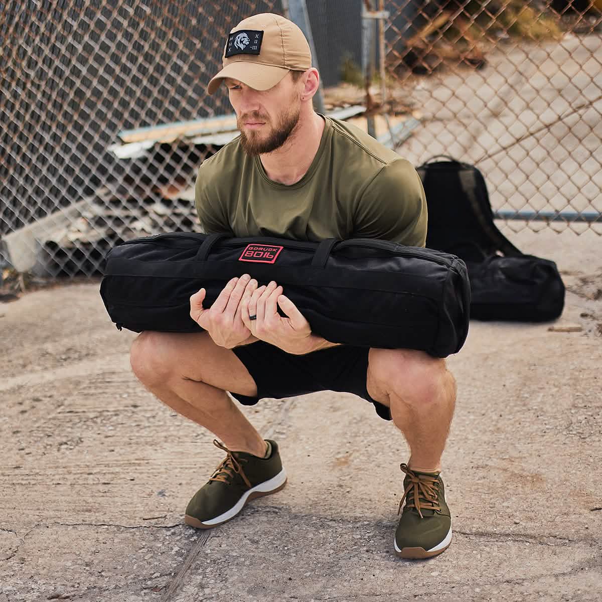 A man wearing a green shirt and cap demonstrates the GORUCK Sandbag Strength with Cadre DS (6-Week Training Program) in front of a chain-link fence. He's outdoors, embracing tactical fitness with his shorts and athletic shoes.