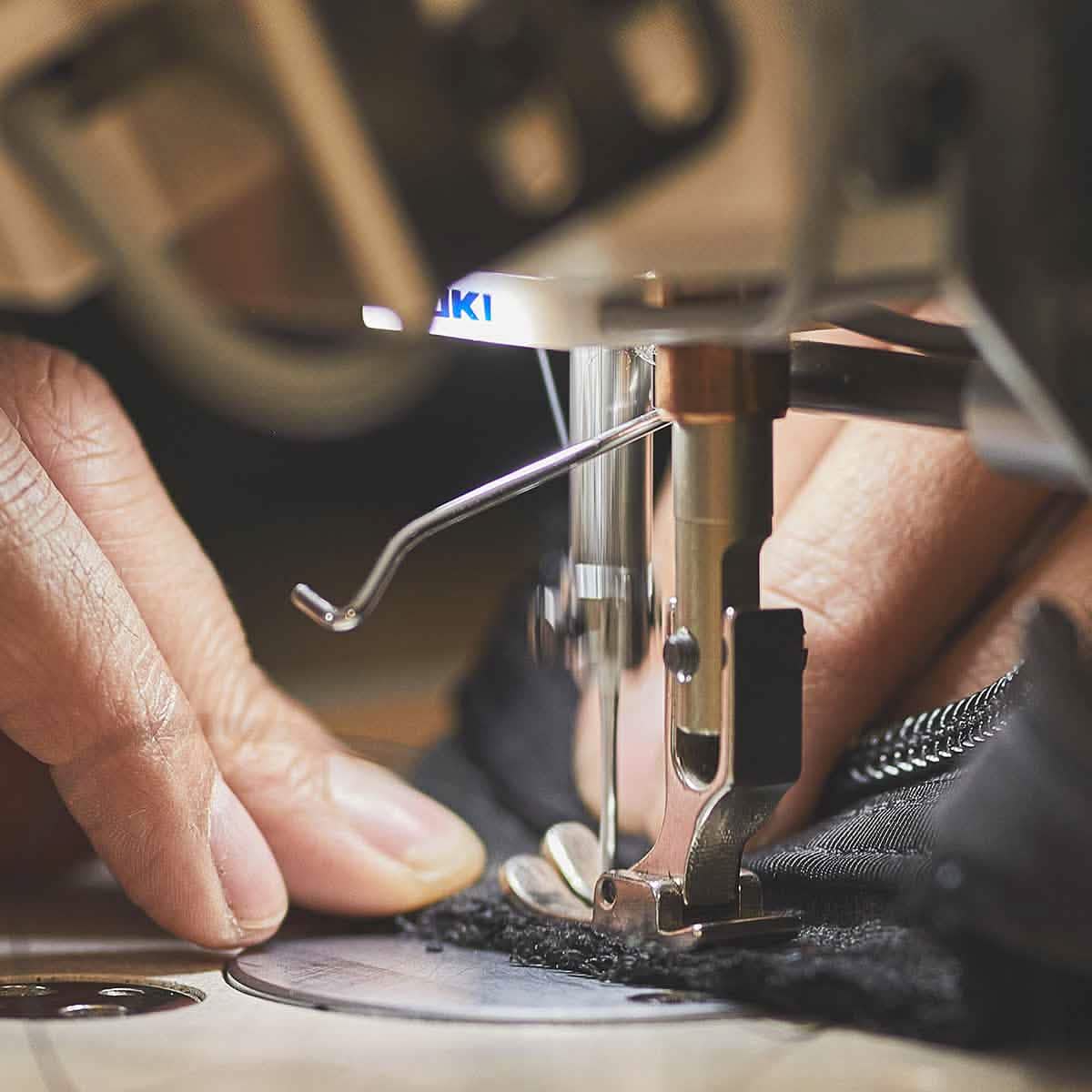 A close-up captures a person's hands skillfully guiding fabric through a sewing machine, the needle stitching with precision as a zipper adorns the black material. This scene reflects the meticulous craftsmanship akin to the Scars Customization Form by GORUCK.