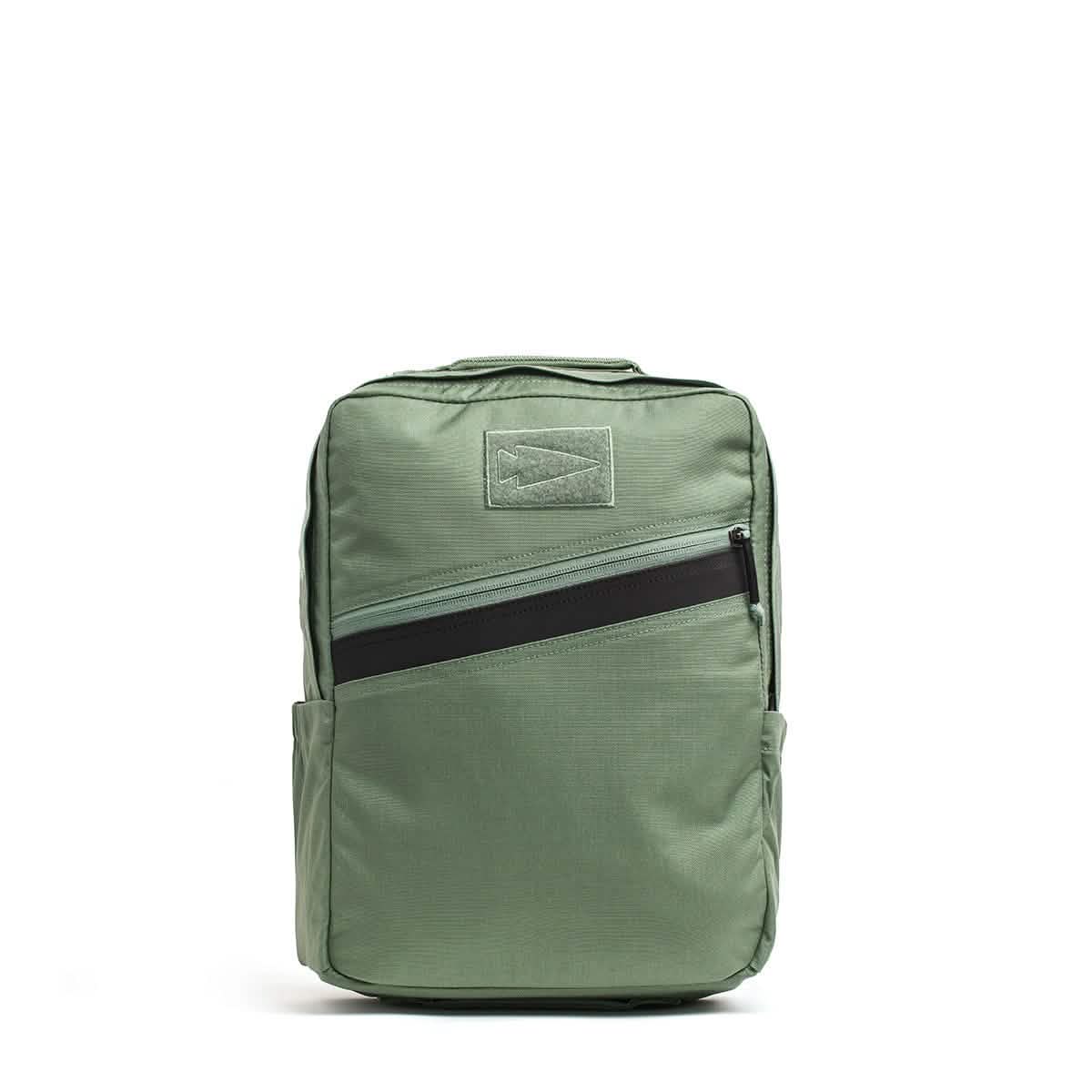 The KR1 by GORUCK is a green kids rucksack with a rectangular shape, featuring a black diagonal zipper pocket on the front. It includes a top handle, padded straps, and side pockets. Its simple design makes it suitable for both school supplies or professional use.