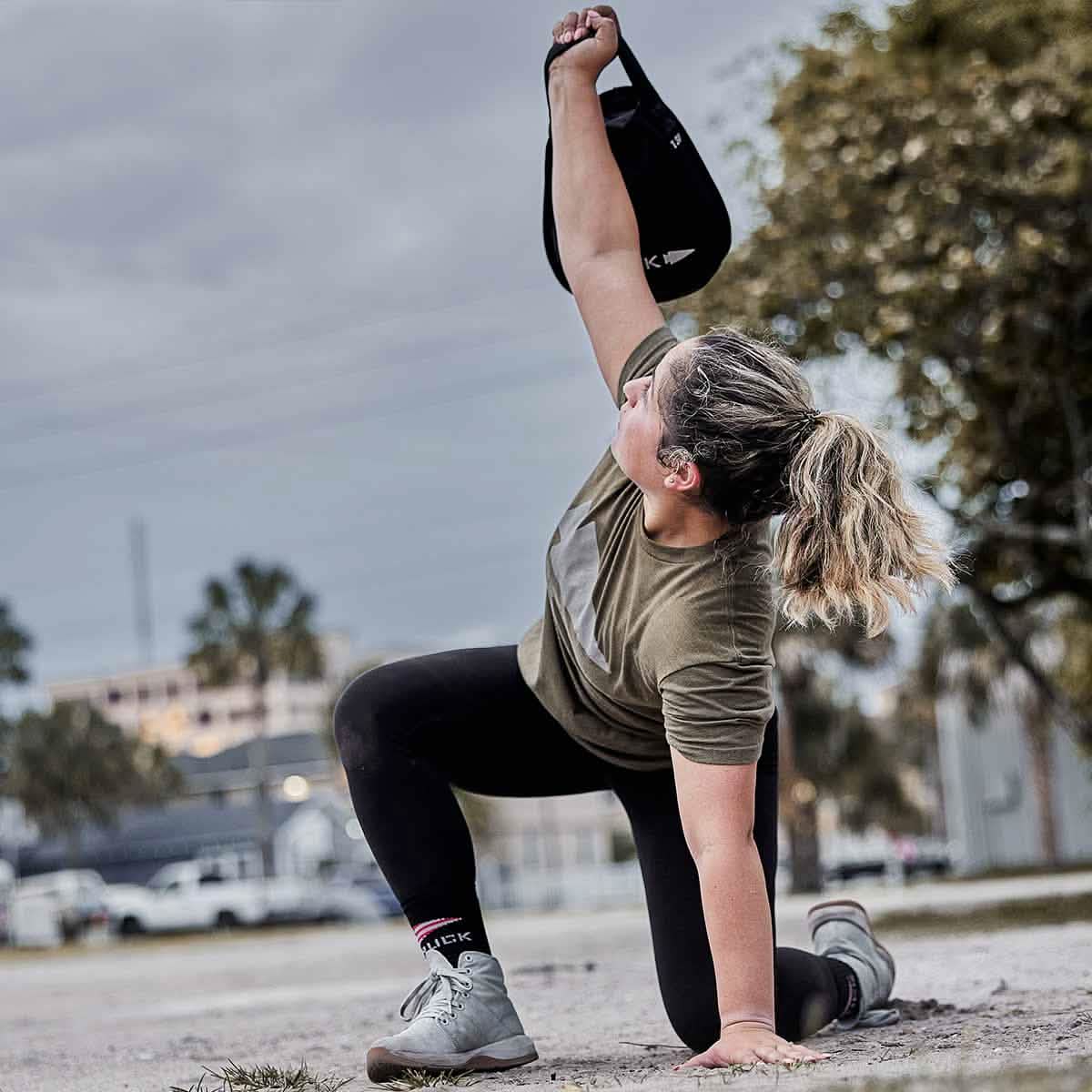 A person with blonde hair engages in an outdoor workout on a sandy surface, lifting a GORUCK Sand Kettlebell overhead with one hand. Wearing a green shirt and black leggings, they exercise under a cloudy sky, surrounded by trees and buildings.