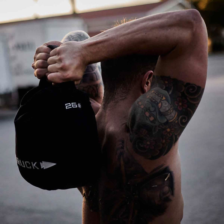 A person with tattooed arms lifts a GORUCK Sand Kettlebell over their shoulder during an outdoor workout, showcasing strength and focus in its endurance-driven design.