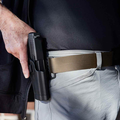 A person wearing a black shirt and light pants is holding a handgun secured in a Spearhead Tactical Belt's black holster by Grip 6, positioned on their right hip.