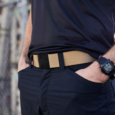 A person wearing a black shirt and pants with hands in pockets, drawing attention to a beige Spearhead Tactical Belt by Grip 6 around their waist. A black digital wristwatch adorns their wrist, while the slightly blurred background reveals a chain-link fence.