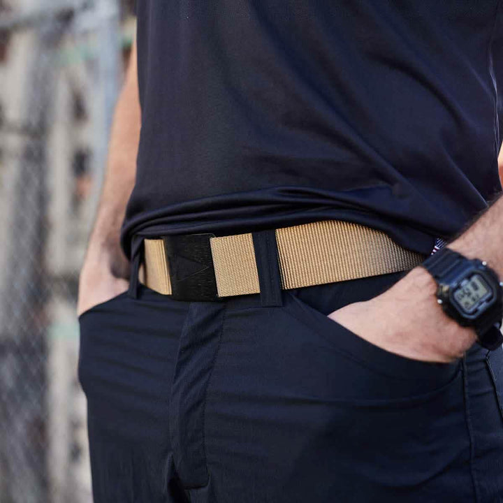 A person wearing a black shirt and pants with hands in pockets, drawing attention to a beige Spearhead Tactical Belt by Grip 6 around their waist. A black digital wristwatch adorns their wrist, while the slightly blurred background reveals a chain-link fence.