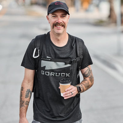 Semi-famous man with a mustache, wearing a black Three Rules Tee - Tri-Blend from GORUCK and a backpack, holding a coffee cup and smiling outdoors.