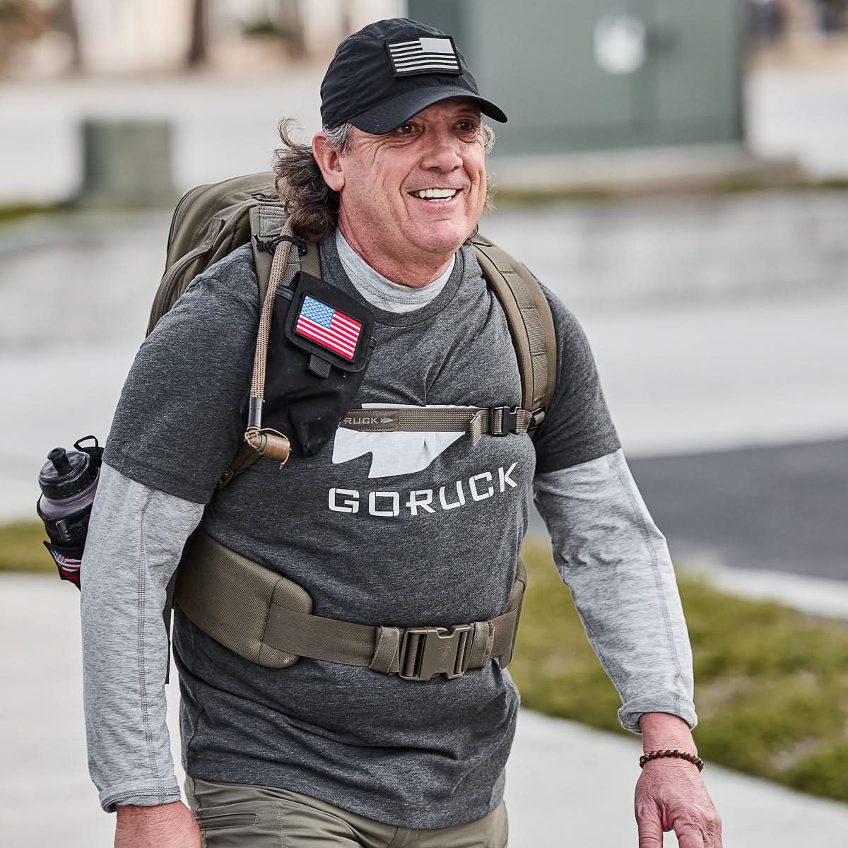 A smiling man outdoors wears the GORUCK Three Rules Tee - Tri-Blend and cap, carrying a backpack with an American flag patch—ideal for standout photos.