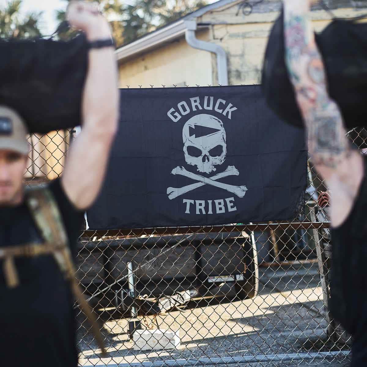 Two people lift weights in front of a Flag - Tribe banner adorned with a skull design, capturing the essence of the GORUCK Challenge.
