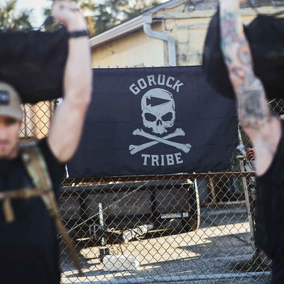 Two people lift weights in front of a Flag - Tribe banner adorned with a skull design, capturing the essence of the GORUCK Challenge.