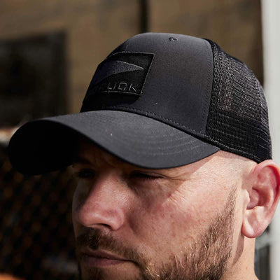 A man is facing forward wearing the GORUCK Performance Trucker Hat - Spearhead, which features a black mesh design and an adjustable back velcro strap. The cap sports a rectangular patch on the front, while the blurred background shows glimpses of a building and a fence.