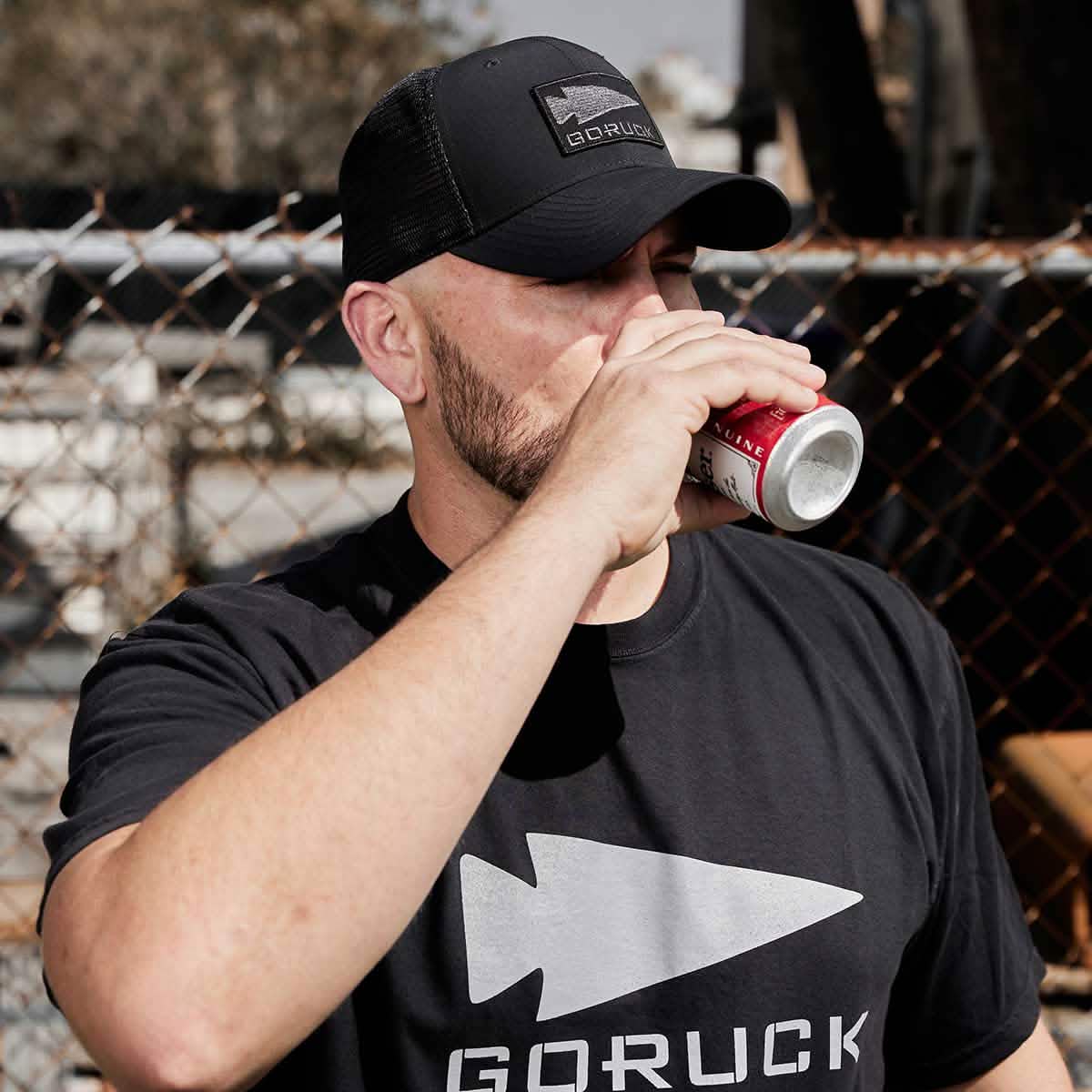 A person wearing a black GORUCK t-shirt and the Performance Trucker Hat - GORUCK Spearhead, which features an adjustable velcro strap, is sipping from a can. They are outdoors in front of a chain-link fence.