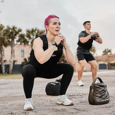 Two people perform squats outdoors. The woman in front, sporting pink hair, wears the GORUCK Training Weight Vest 2.0 in black. Her companion in the background dons a gray outfit. Nearby rest a weighted bag and ball, with palm trees and buildings serving as the backdrop.