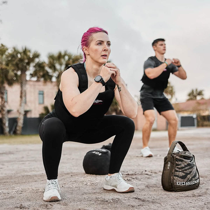 Two people perform squats outdoors. The woman in front, sporting pink hair, wears the GORUCK Training Weight Vest 2.0 in black. Her companion in the background dons a gray outfit. Nearby rest a weighted bag and ball, with palm trees and buildings serving as the backdrop.