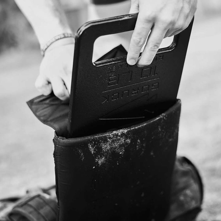 A person places a black 10-pound GORUCK Training Weight Vest 2.0 into a backpack outdoors. The close-up image captures the weight halfway inside the bag, with the person's hands visible, resembling someone loading an adjustable weighted vest. The scene is in black and white.